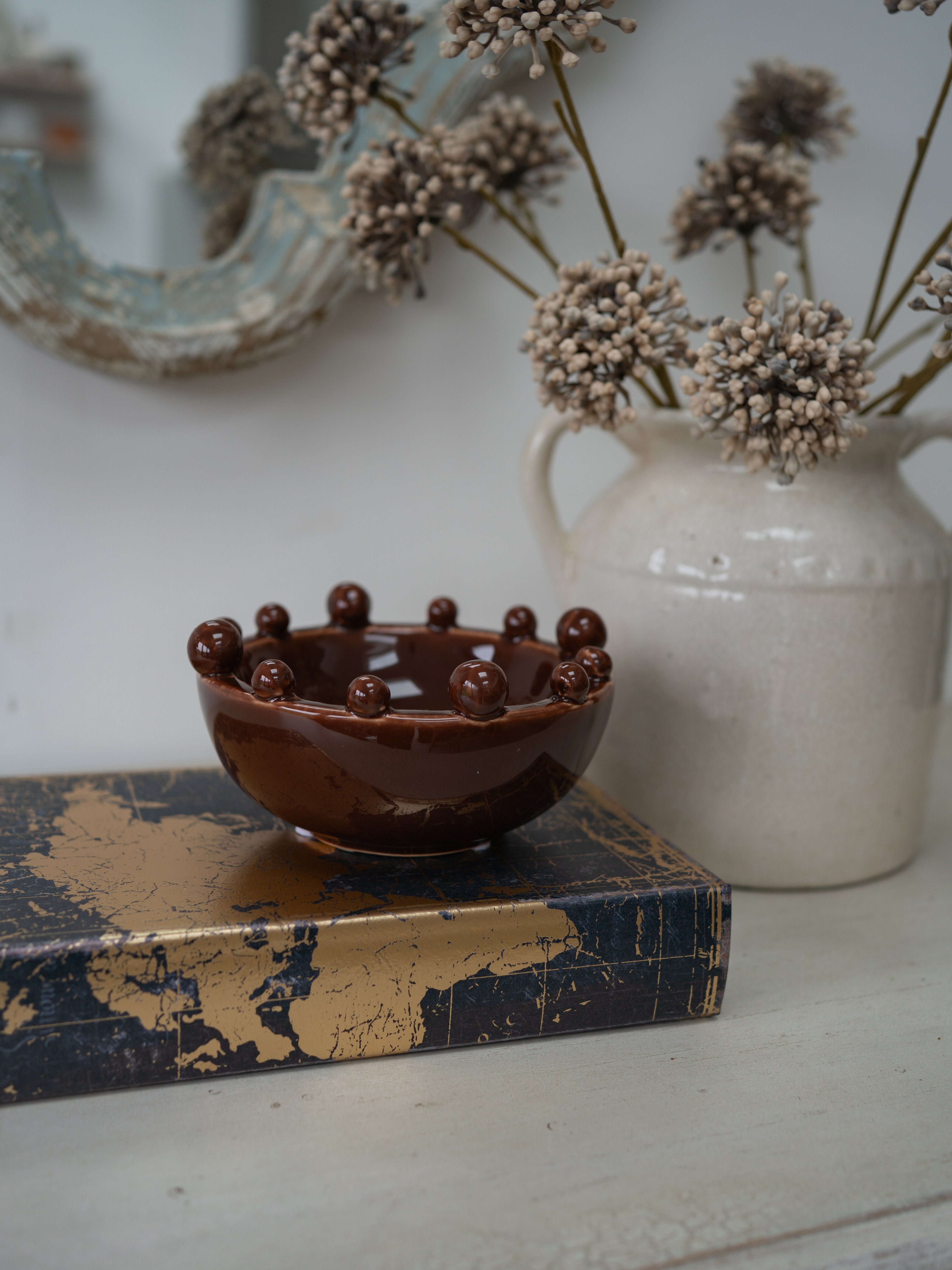A small glossy brown ceramic bowl with raised spherical details around the rim, displayed on a dark blue and gold book in front of a white ceramic vase with dried flowers.