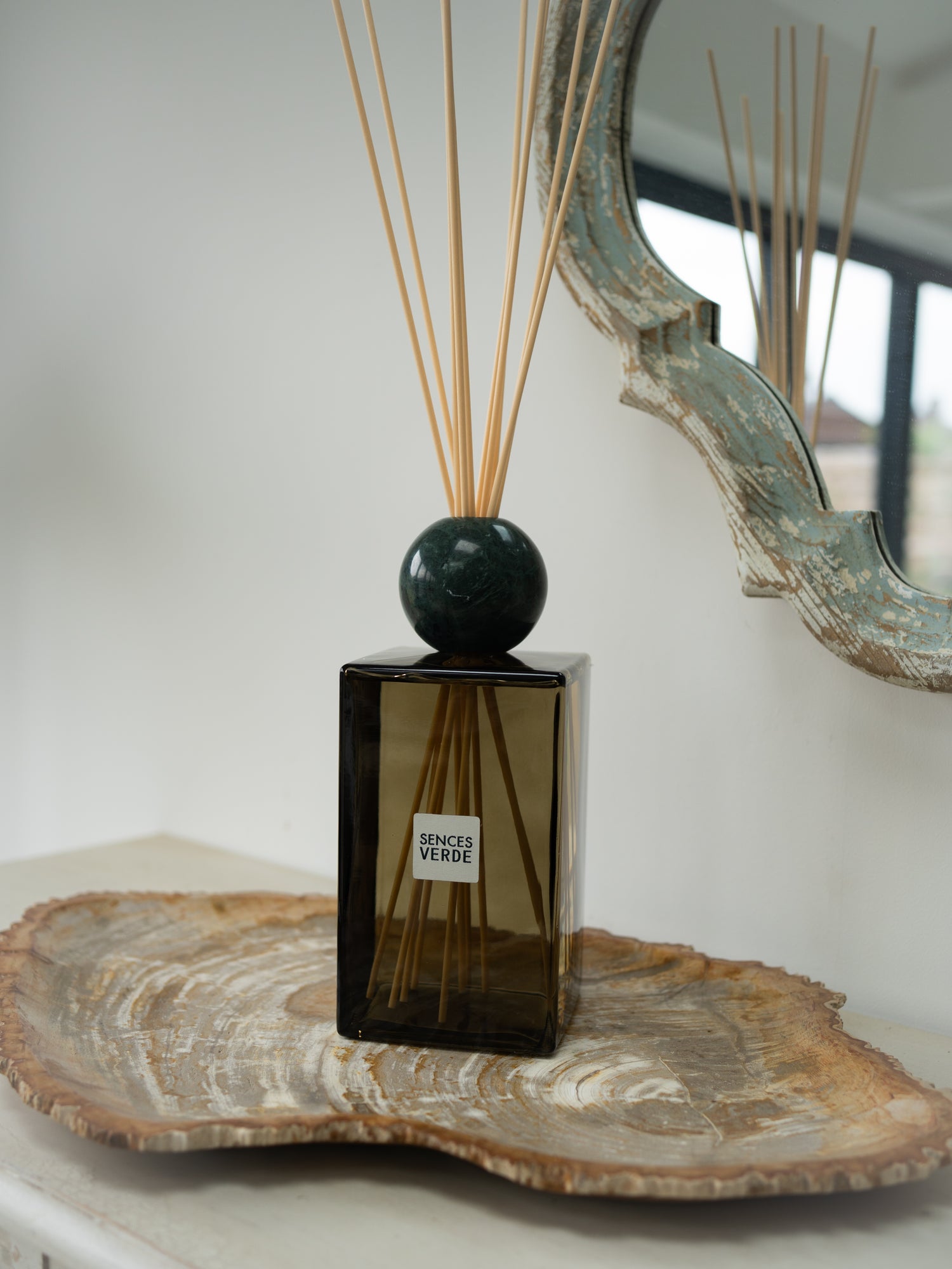 A reed diffuser bottle with a dark green glass body, a round dark green marble top, and natural reed sticks, styled on a wooden tray in a home interior setting.