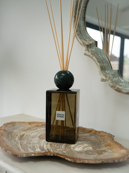 A reed diffuser bottle with a dark green glass body, a round dark green marble top, and natural reed sticks, styled on a wooden tray in a home interior setting.