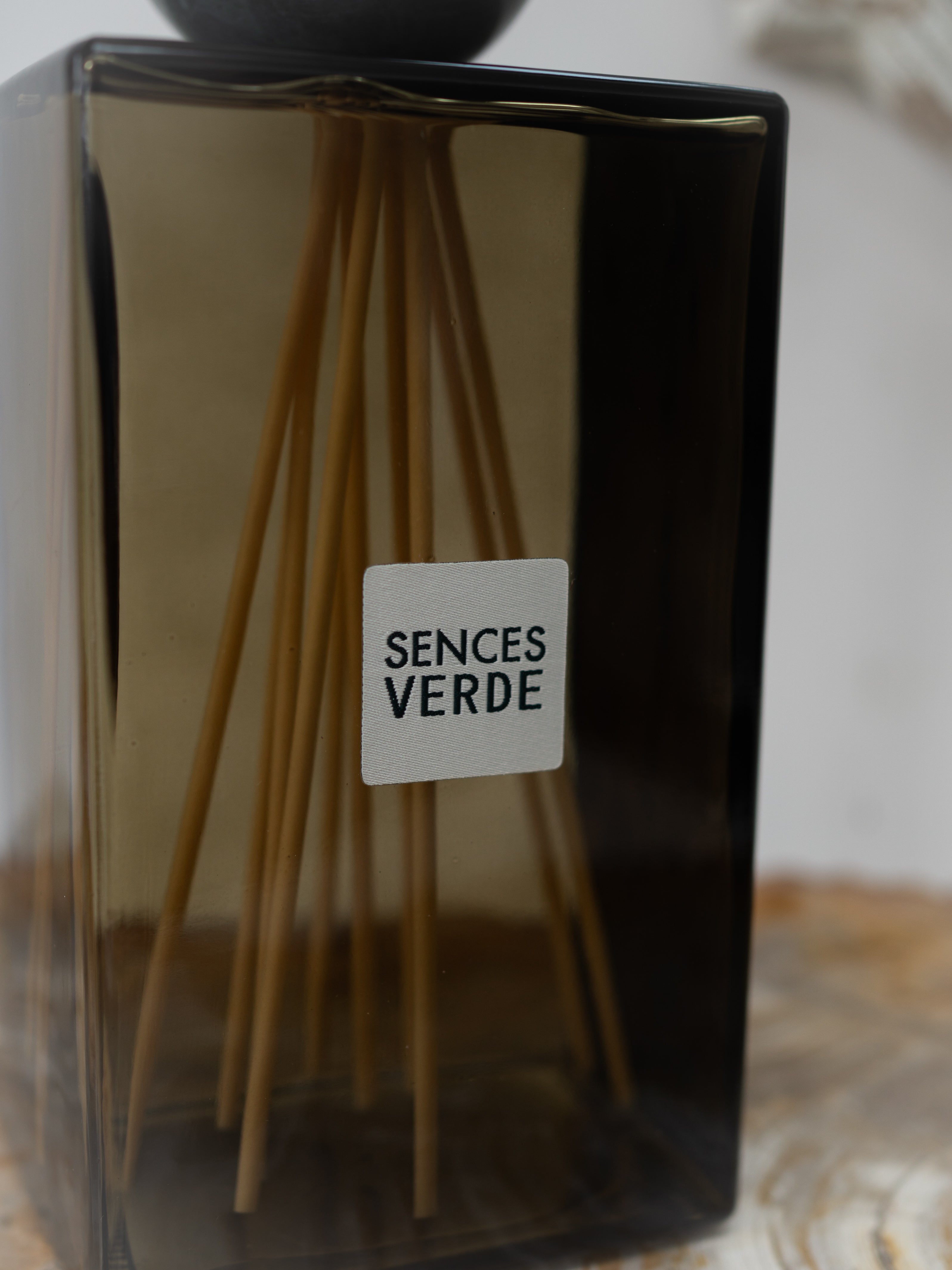 A reed diffuser bottle with a dark green glass body, natural wood reeds, and a white label reading &