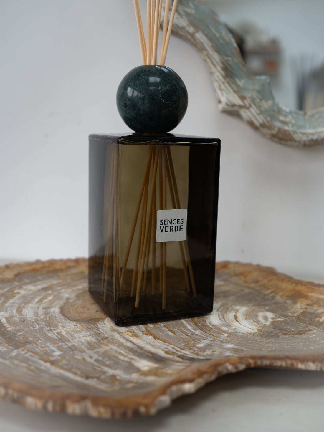 A reed diffuser bottle made of dark green glass with a round dark green marble top and natural reeds, sitting on a rustic wood slice tray. The bottle has a white label that reads &