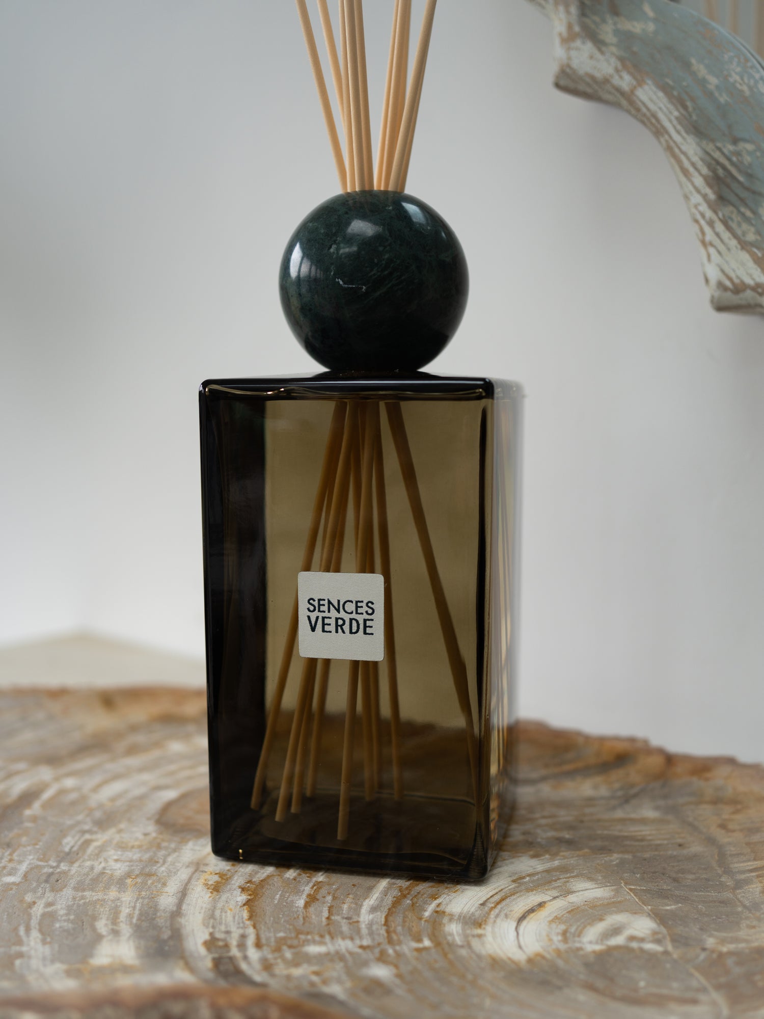 A Sences Verde reed diffuser with a dark green glass bottle, a polished dark green marble sphere, and natural reed sticks, displayed on a rustic wood slice.