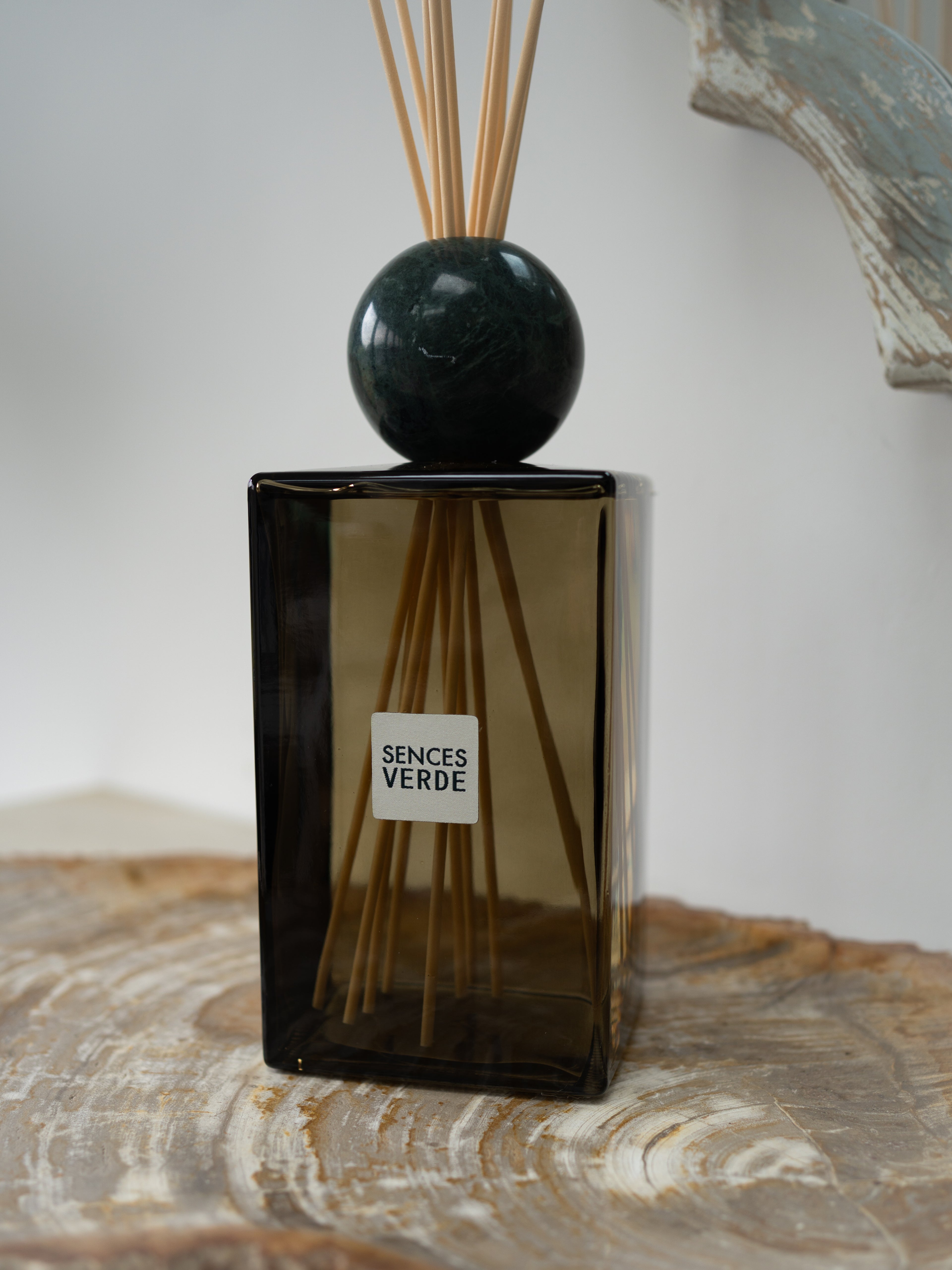 A Sences Verde reed diffuser with a dark green glass bottle, a polished dark green marble sphere, and natural reed sticks, displayed on a rustic wood slice.