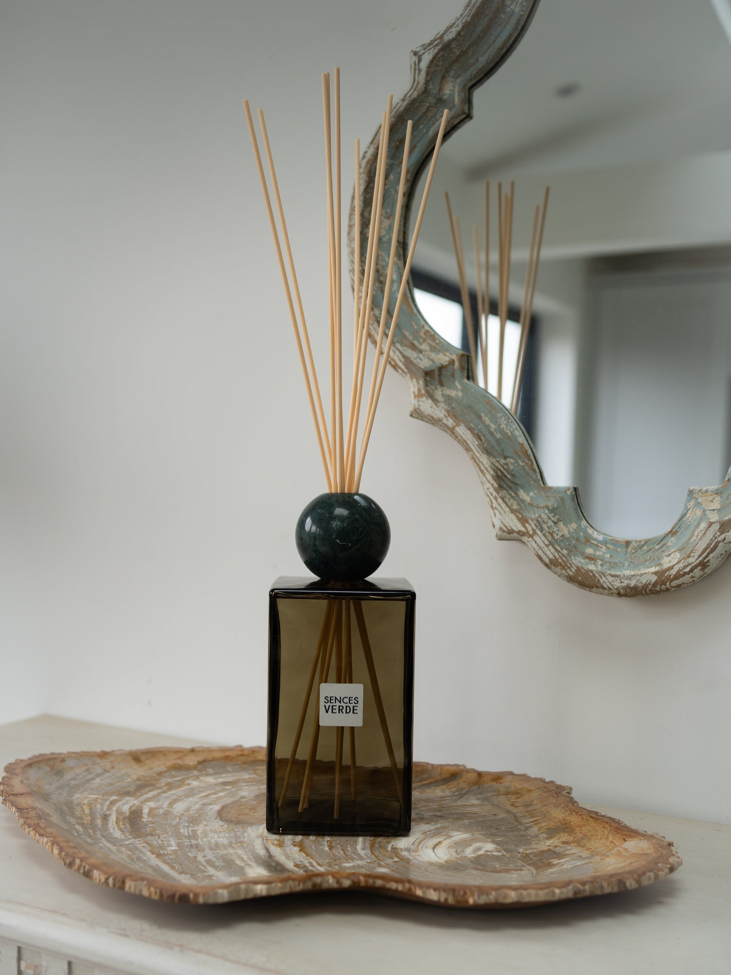 A reed diffuser with a dark green glass bottle, a round green marble stopper, and natural reed sticks, displayed on a rustic wood slice tray against a neutral wall with a decorative mirror.