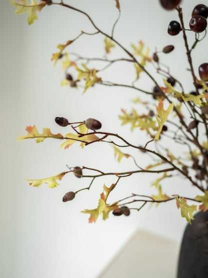 Autumn artificial branch arrangement with yellow oak leaves, brown acorns, and dark berries on thin branches, photographed against a soft light background.