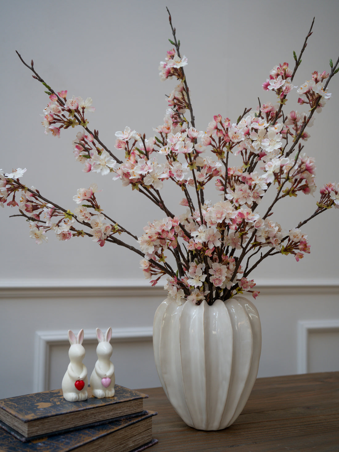 Vase with cherry blossom branches on a table with books and rabbit figurines.