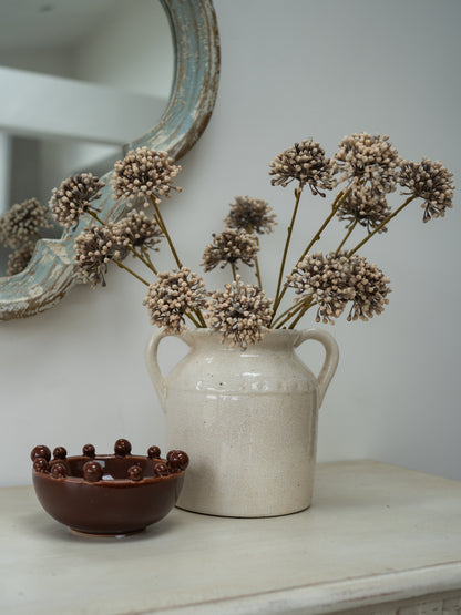 A rustic cream-colored ceramic vase filled with faux beige allium flower clusters, placed on a light tabletop next to a small dark brown ceramic bowl, with a distressed mirror visible in the background.