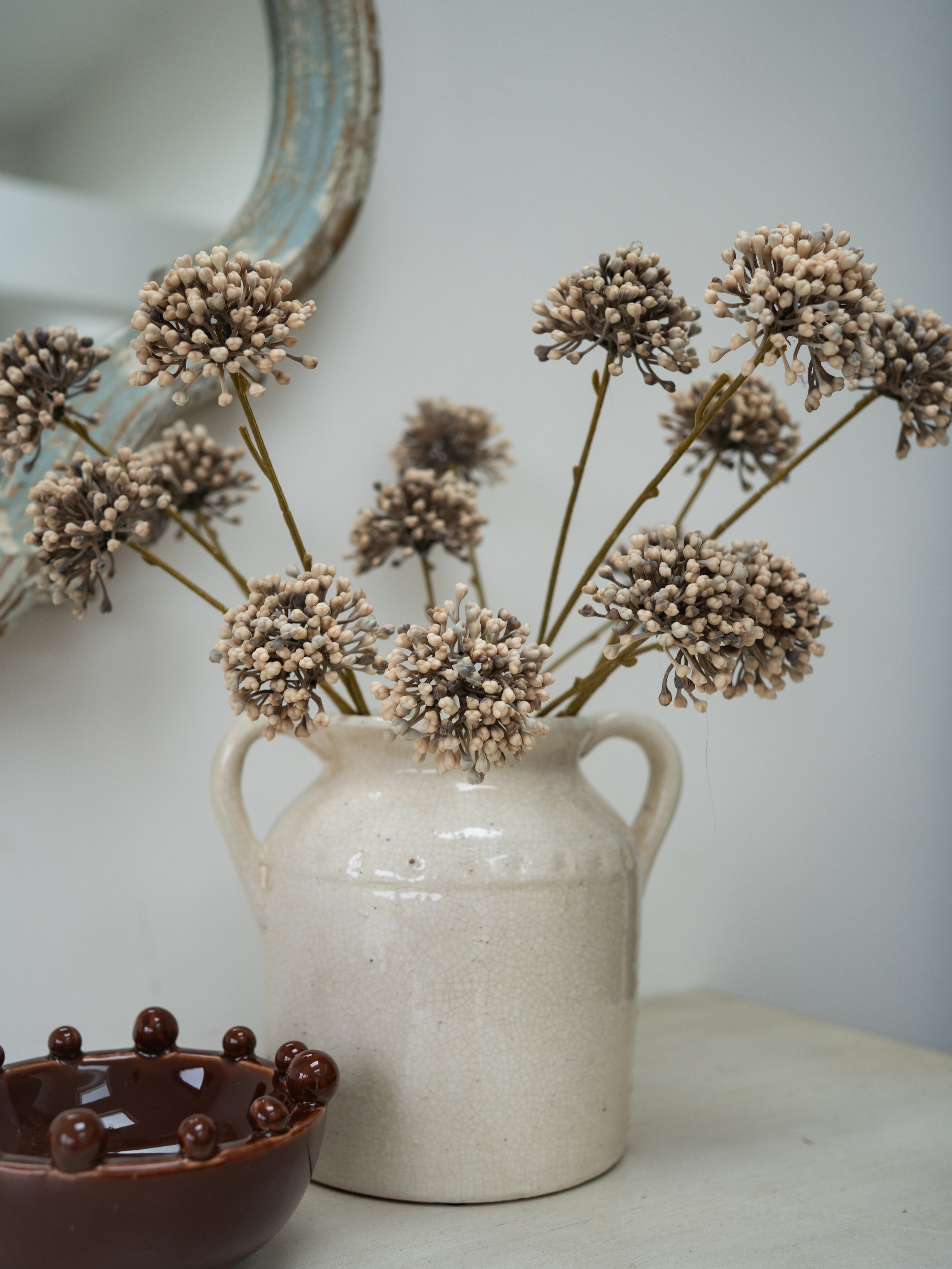 A rustic cream-colored ceramic pitcher vase holding dried beige allium flower stems, styled on a tabletop with a dark brown bowl in the foreground and a mirror edge in the background.