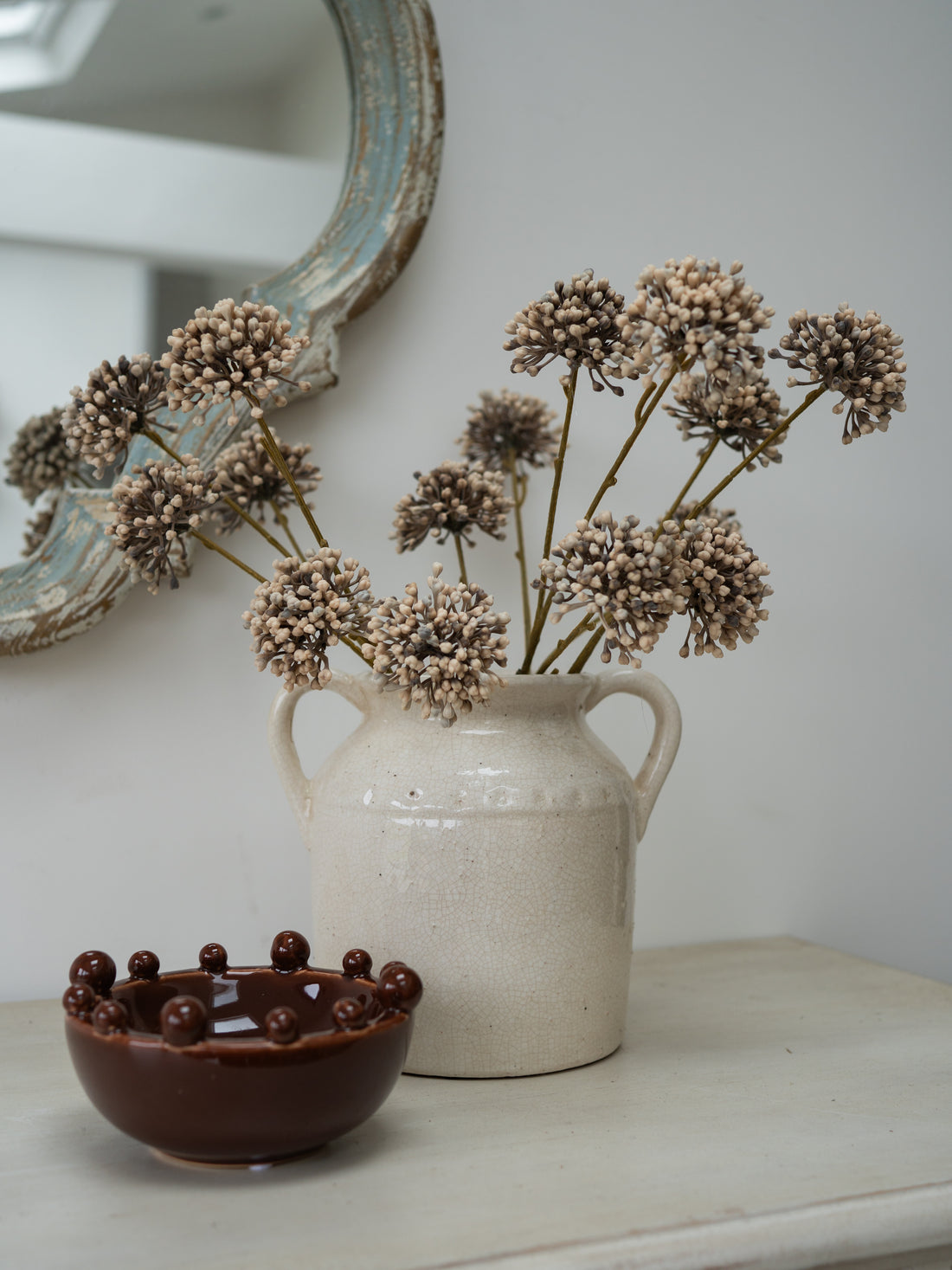 A rustic cream-colored ceramic pitcher vase filled with dried beige allium, styled on a light tabletop next to a small dark brown ceramic bowl with raised bead details, with a distressed mirror in the background.