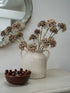 A rustic cream-colored ceramic pitcher vase filled with dried beige allium, styled on a light tabletop next to a small dark brown ceramic bowl with raised bead details, with a distressed mirror in the background.