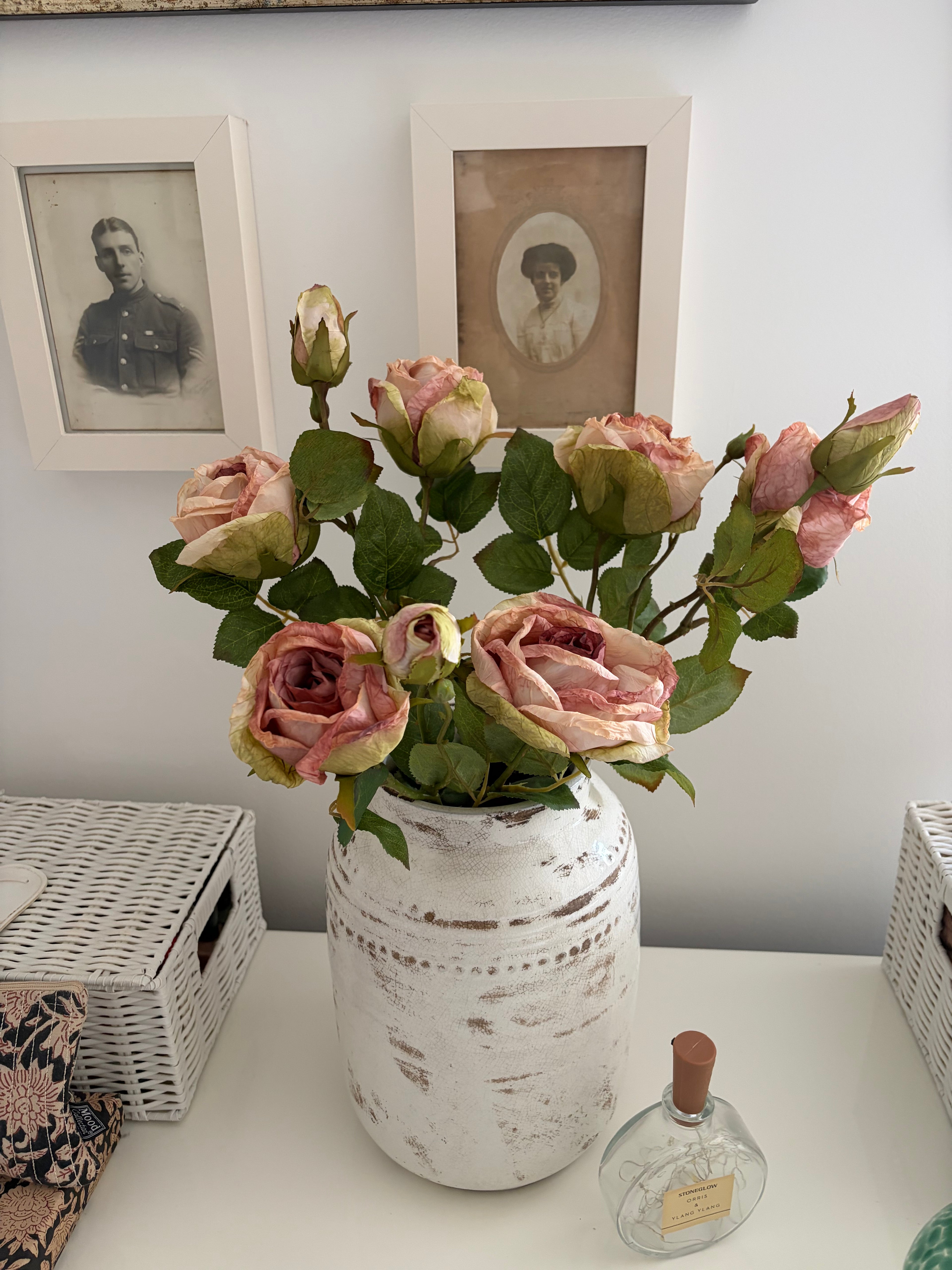 A bouquet of artificial pink roses with green leaves arranged in a distressed white ceramic vase, placed on a white table with framed vintage photos and wicker baskets in the background.