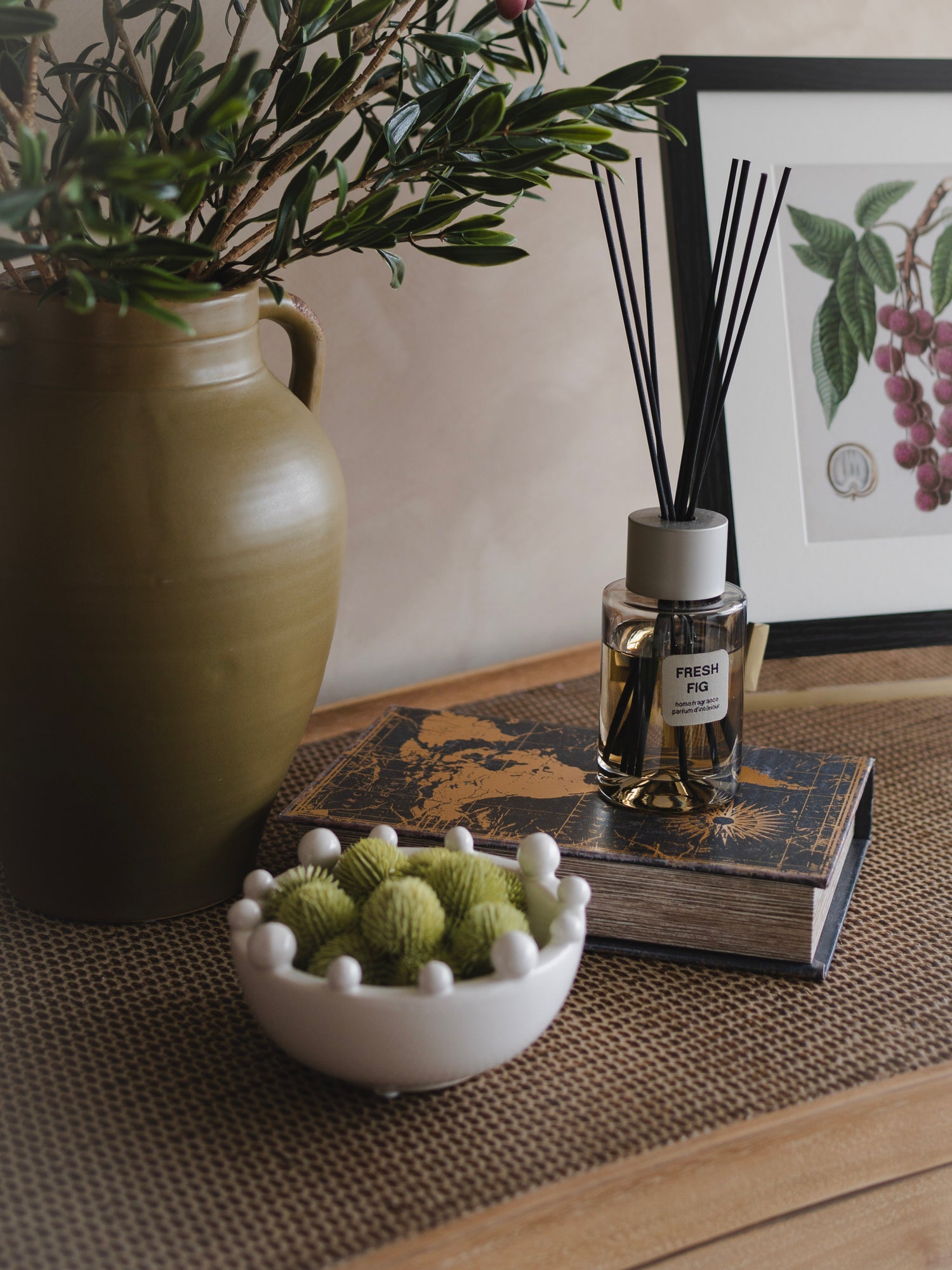 small white ceramic bowl with bobbles, filled with faux green echinops and fresh fig luxury reed diffuser with green vase filled with faux olive stems in the background