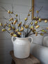 A rustic white ceramic jugsard vase filled with dried branches featuring small green and tan acorns, displayed on a wooden stool in a cosy living room setting.