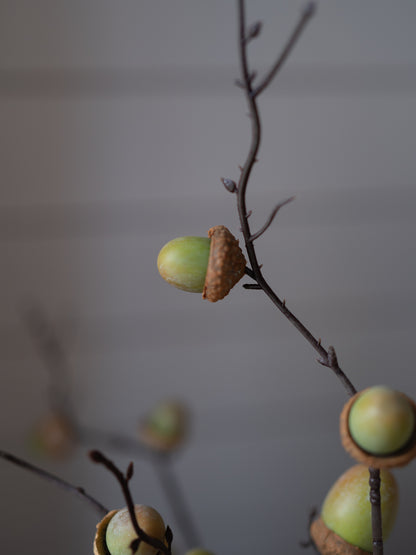 Faux acorns with brown textured caps attached to thin brown branches, arranged as a decorative garland against a soft, blurred gray background.