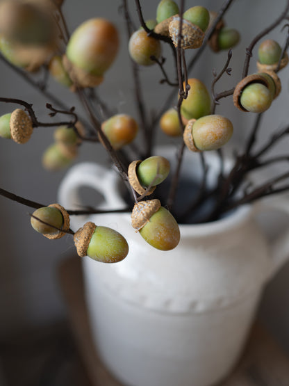 Faux acorn branch decor with green and tan acorns on dark brown branches, arranged in a white ceramic pitcher against a soft gray background.