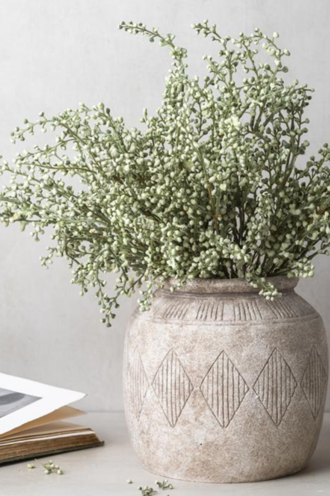 A textured beige ceramic vase with a carved diamond pattern, filled with faux green heather stem, set against a neutral light gray background.