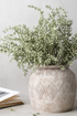 A textured beige ceramic vase with a carved diamond pattern, filled with faux green heather stem, set against a neutral light gray background.