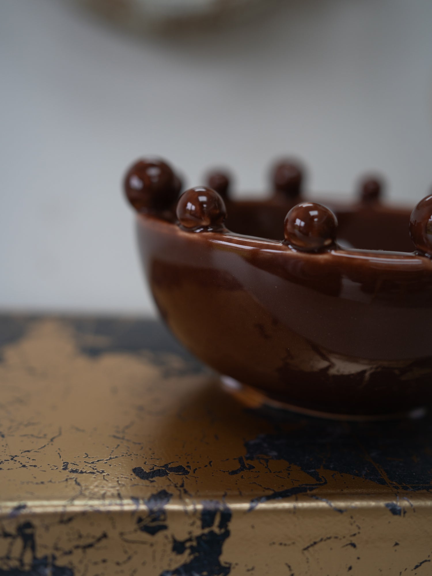 A close-up of a glossy dark brown ceramic bowl with raised spherical details around the rim, resting on a distressed gold and navy surface against a soft blurred light gray background.