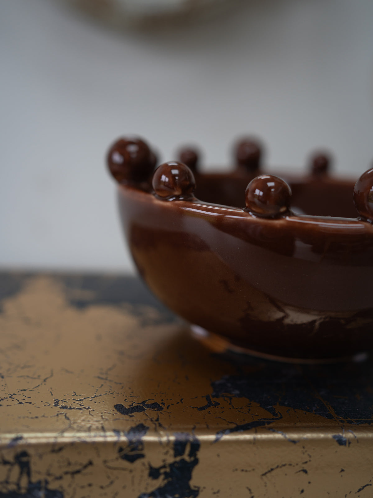 A close-up of a glossy dark brown ceramic bowl with raised spherical details around the rim, resting on a distressed gold and navy surface against a soft blurred light gray background.