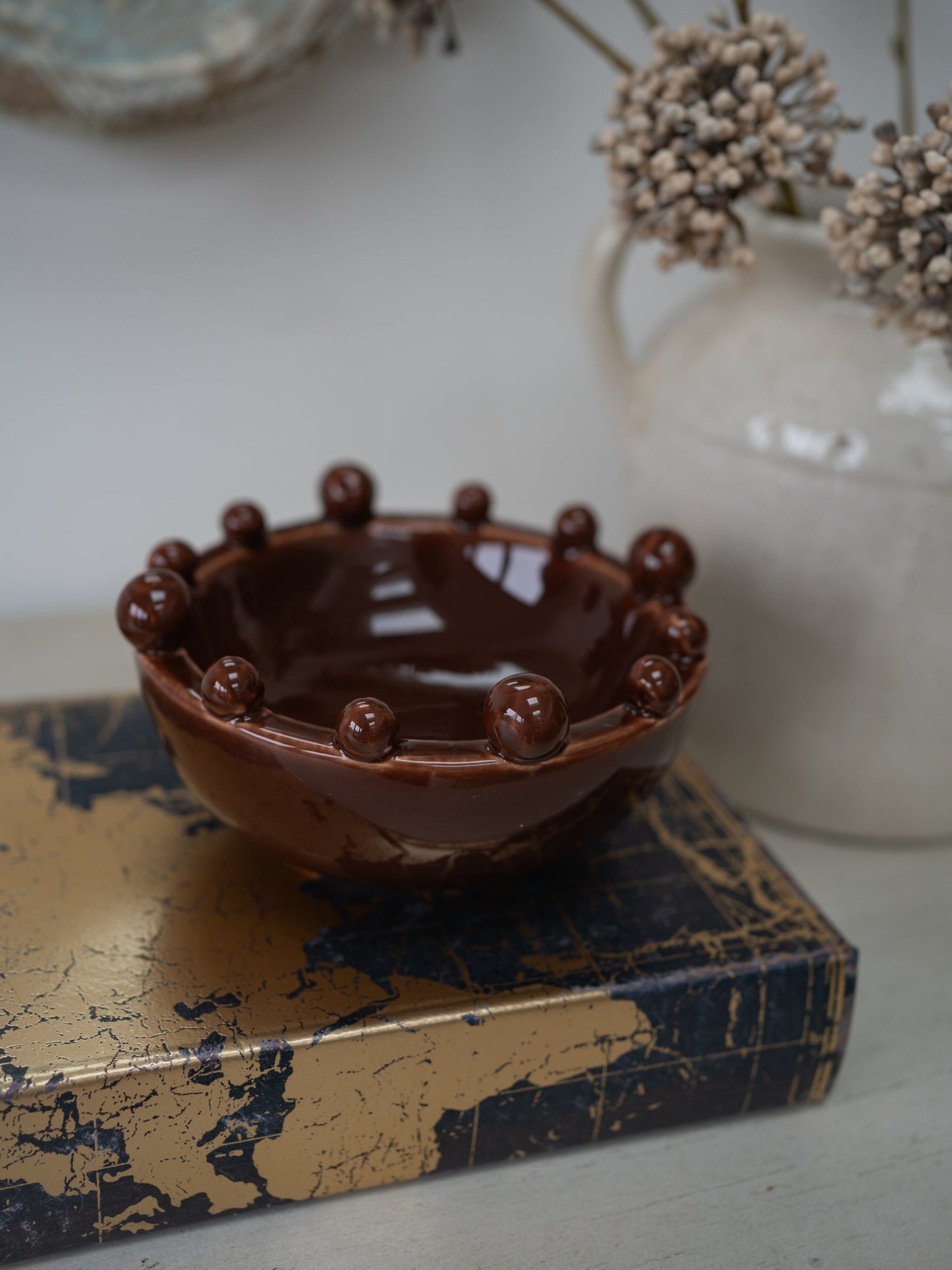 A glossy dark brown ceramic bowl with a raised beaded rim, displayed on a distressed gold and navy book cover with a white vase of dried flowers in the background.