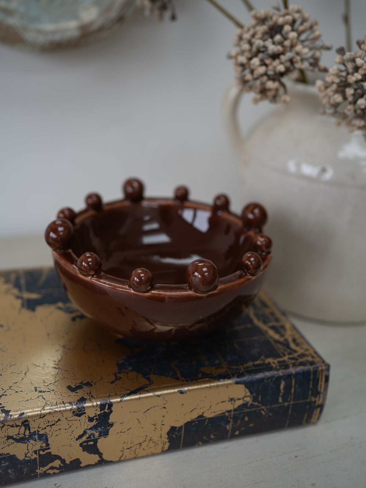 A glossy dark brown ceramic bowl with a raised beaded rim, displayed on a distressed gold and navy book cover with a white vase of dried flowers in the background.