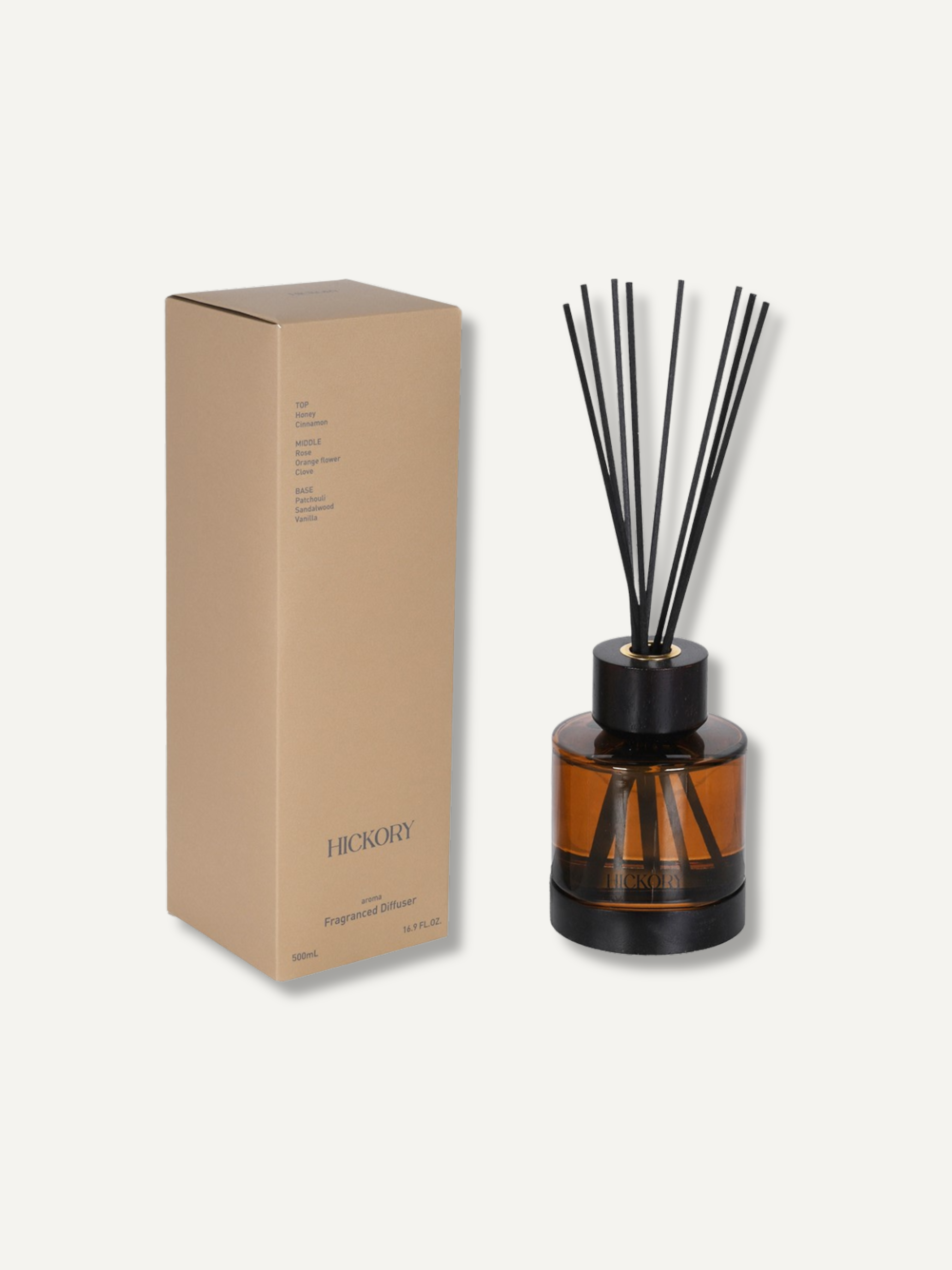 An amber glass reed diffuser bottle with black reeds and a black cap, shown next to its tan cardboard packaging box on a light off-white background. The box is labeled &