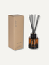An amber glass reed diffuser bottle with black reeds and a black cap, shown next to its tan cardboard packaging box on a light off-white background. The box is labeled &