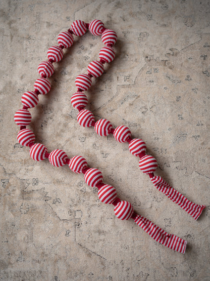 A festive christmas garland made of red and white striped fabric beads, laid out on a textured beige stone surface.