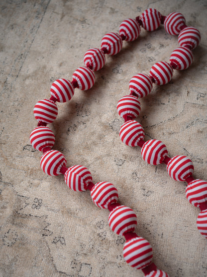 A beaded garland made of fabric, round beads with alternating red and white horizontal stripes, laid out on a textured beige fabric background.