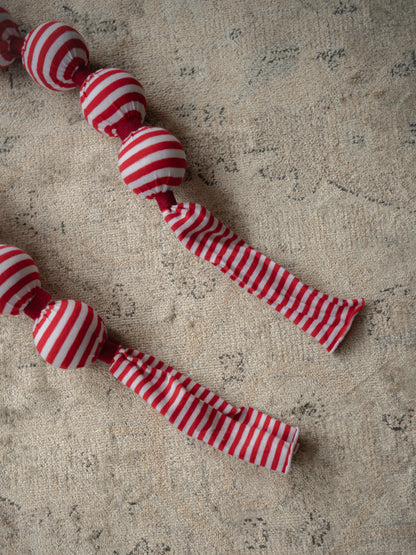 A red and white striped fabric christmas garland with round beads and long ribbon tails, laid diagonally on a textured beige rug.