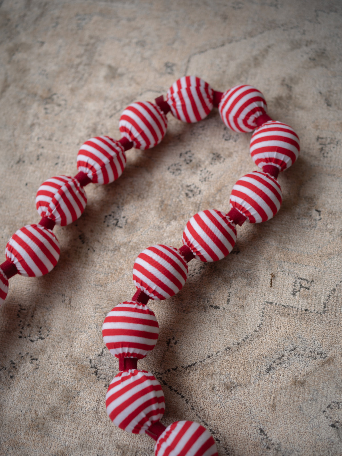 A beaded garland made of round fabric beads with red and white horizontal stripes, laid out on a textured beige surface.
