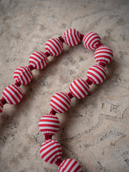 A beaded garland made of round fabric beads with red and white horizontal stripes, laid out on a textured beige surface.