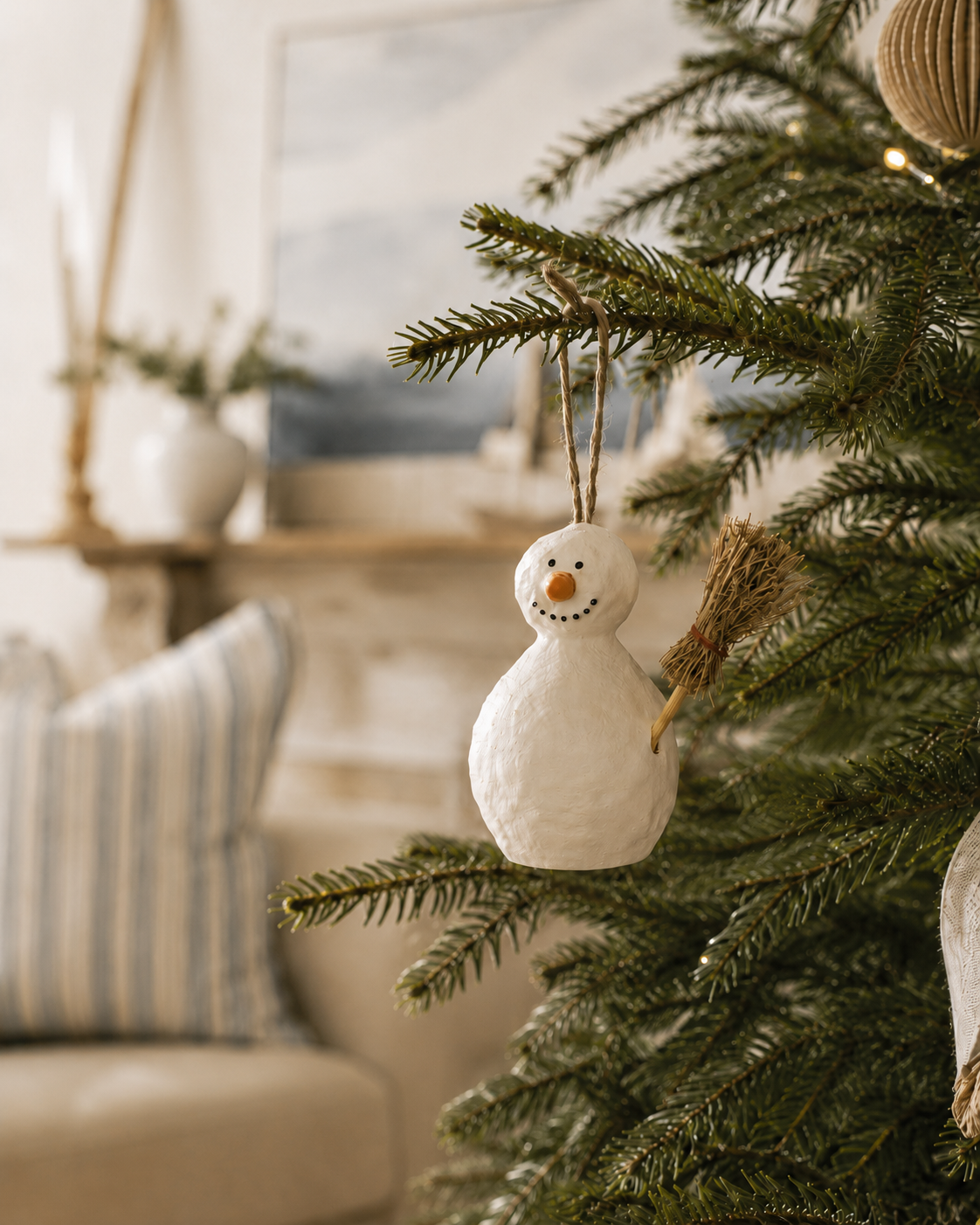 A white textured snowman Christmas ornament with a small straw-colored broom hangs on the branches of a green Christmas tree in a softly lit living room.