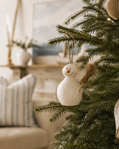 A white textured snowman Christmas ornament with a small straw-colored broom hangs on the branches of a green Christmas tree in a softly lit living room.