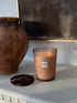 A Scent of Rosso candle in a translucent dusty rose glass jar with a white label, lit and set on a white mantelpiece next to a dark round coaster, with a large brown ceramic pot and dark framed artwork in the background.