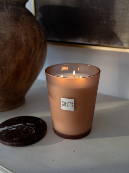 A lit Sences Rosso candle in a frosted amber glass jar, placed on a light gray surface next to a dark round coaster, with a ceramic vase and framed artwork in the background.