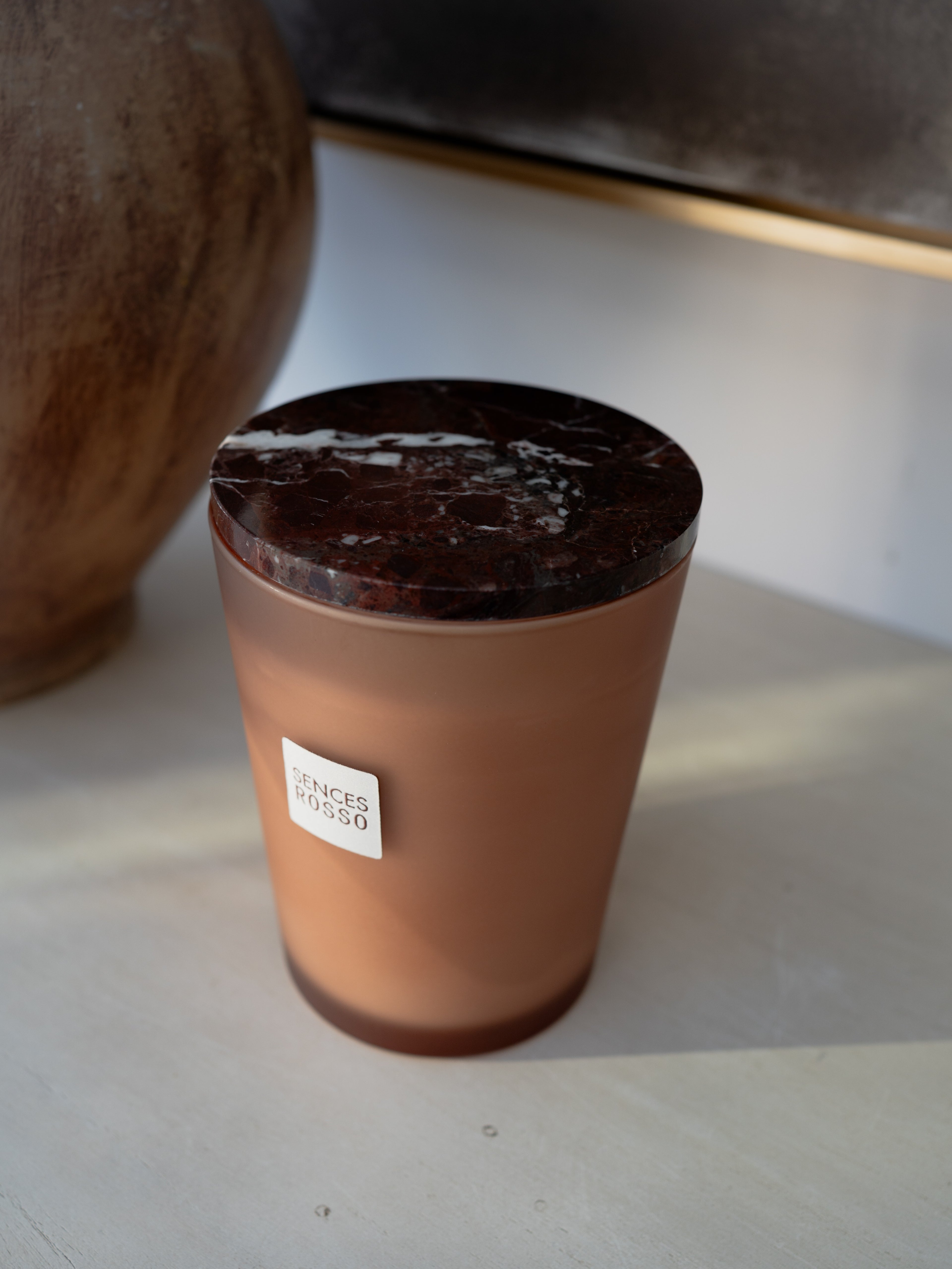 A candle in a frosted terracotta-colored glass jar with a dark brown marble-patterned lid, featuring a small white label that reads &