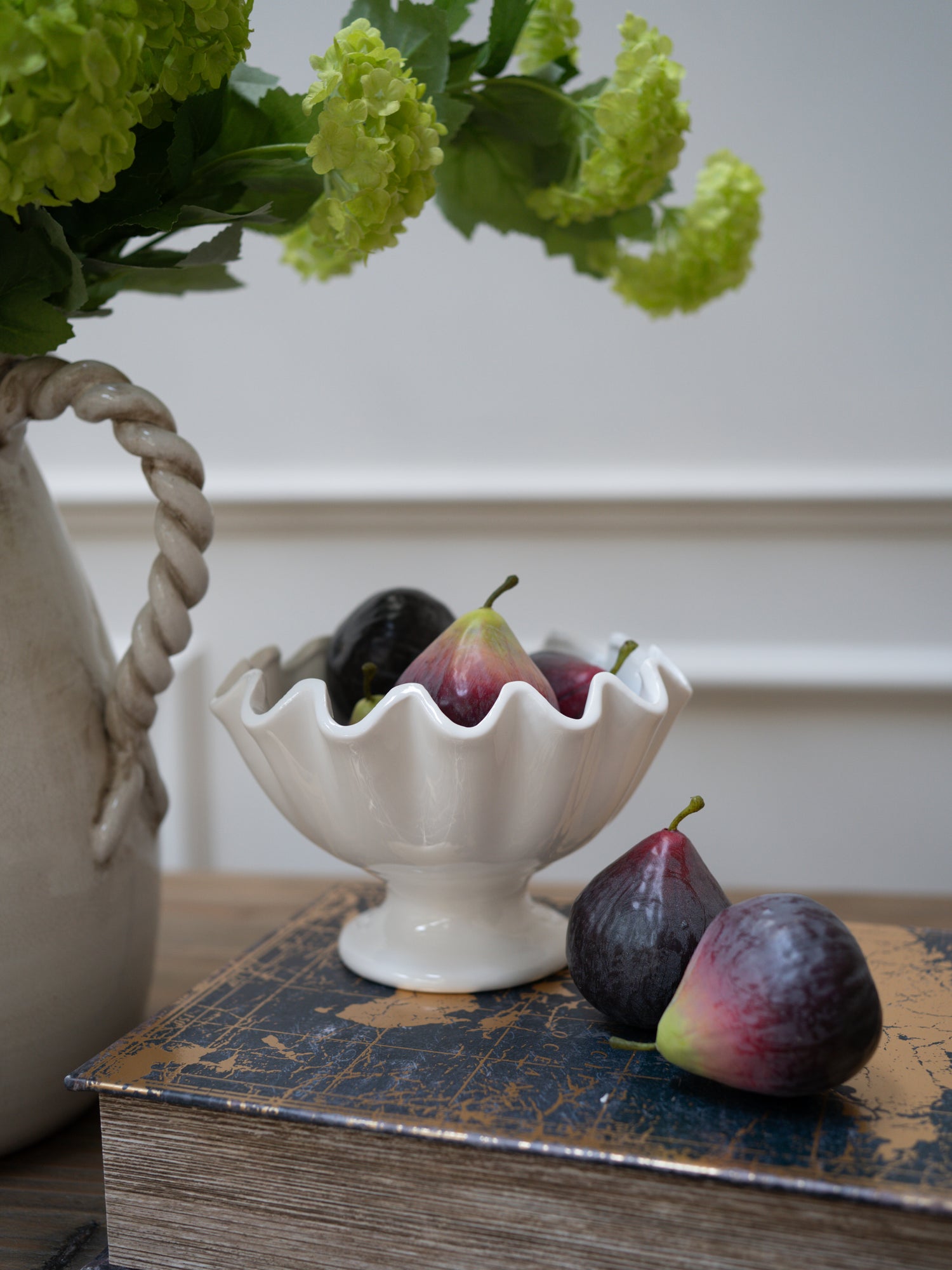 White decorative bowl with faux figs on a wooden surface next to a vase with green flowers. Home styling inspiration and decor ideas
