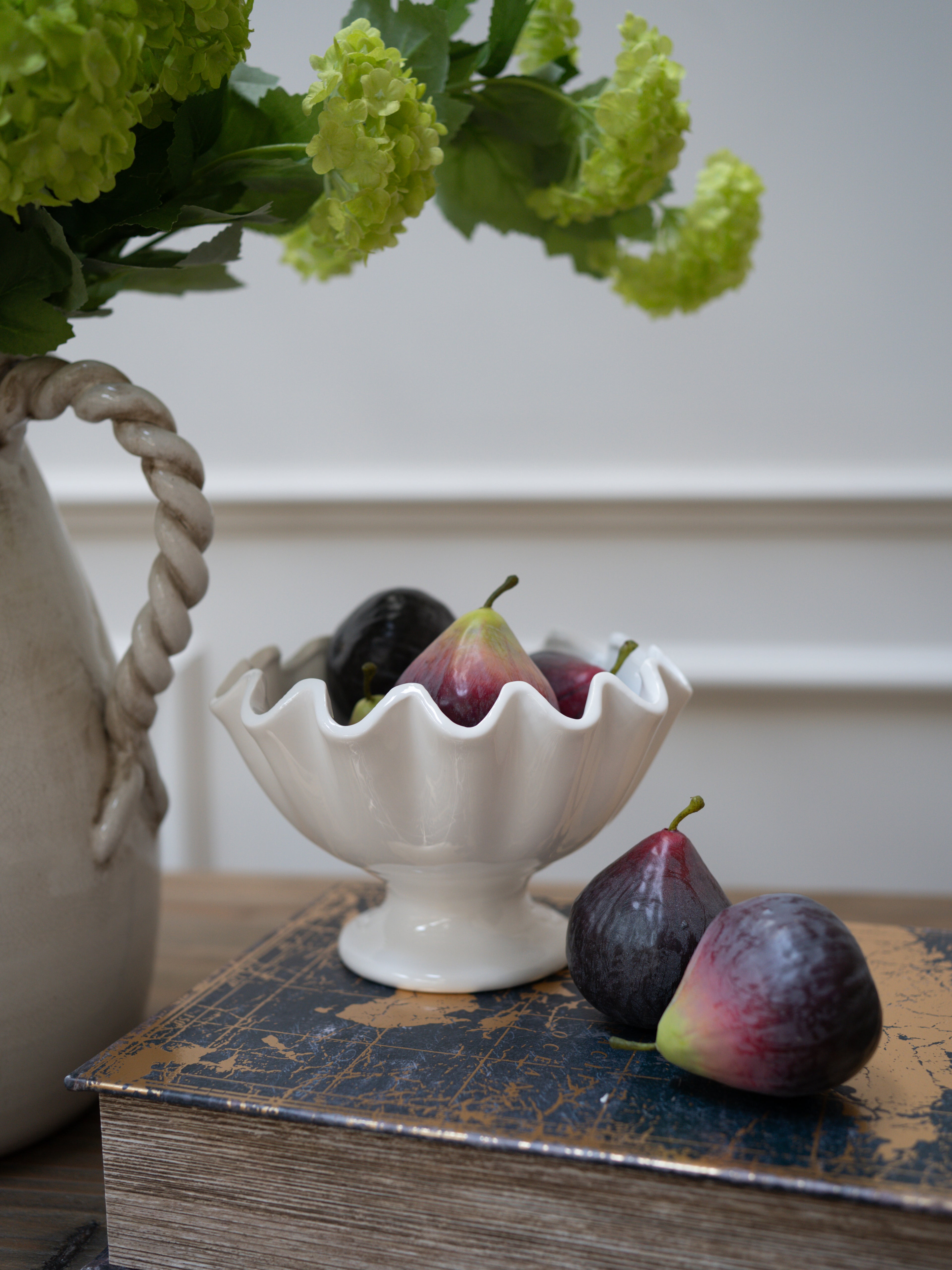 White decorative bowl with faux figs on a wooden surface next to a vase with green flowers. Home styling inspiration and decor ideas