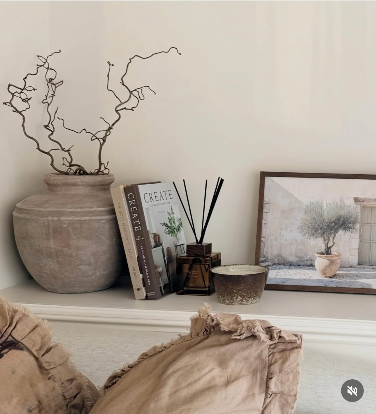 A styled interior shelf scene with a large matte rustic brown ceramic vase holding bare branches, a stack of books including one titled CREATE, a reed diffuser, a small candle, and a framed print against a warm off-white wall.