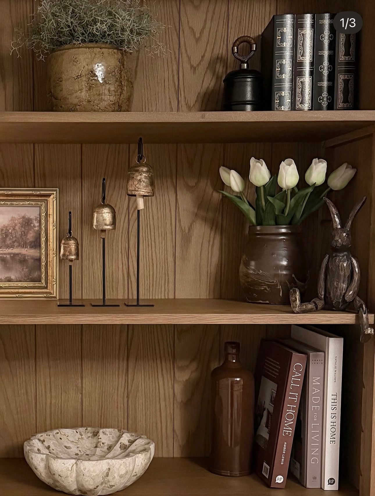 A styled wooden bookshelf with multiple shelves, decorated with a potted plant, books, small brass bell stands, white tulips in a rustic vase, a rabbit figurine, a bowl, and a framed picture.