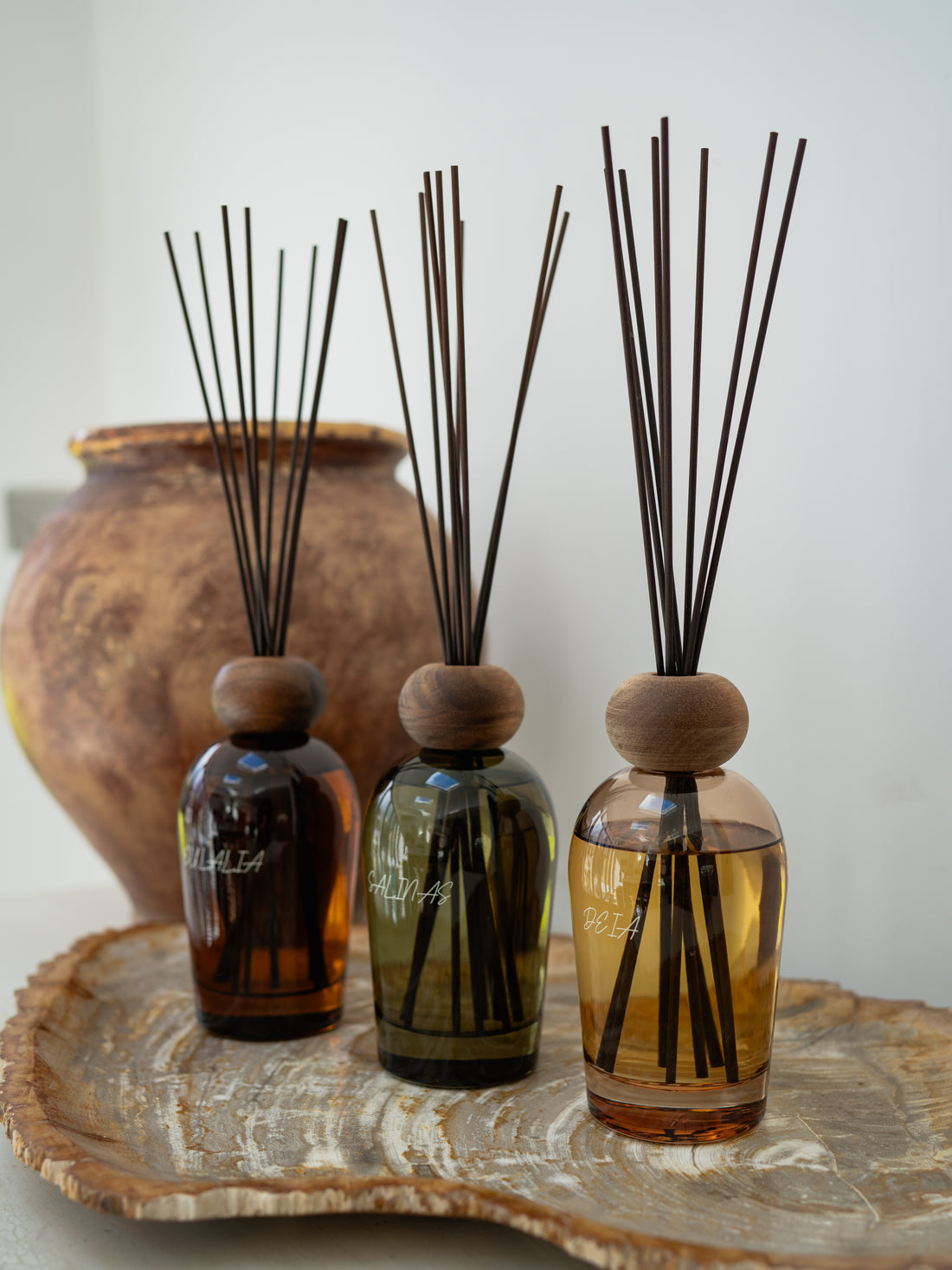 Three balearic inspired diffuser bottles with reeds on a wooden tray against a white background