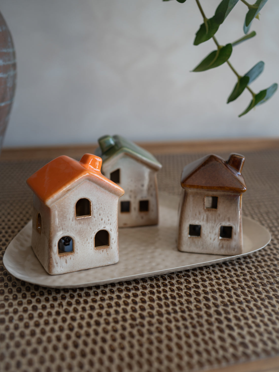 Three small ceramic house-shaped LED holders or decorative figurines in beige, orange, green, and brown glazes, arranged on a matching beige tray over a woven surface.