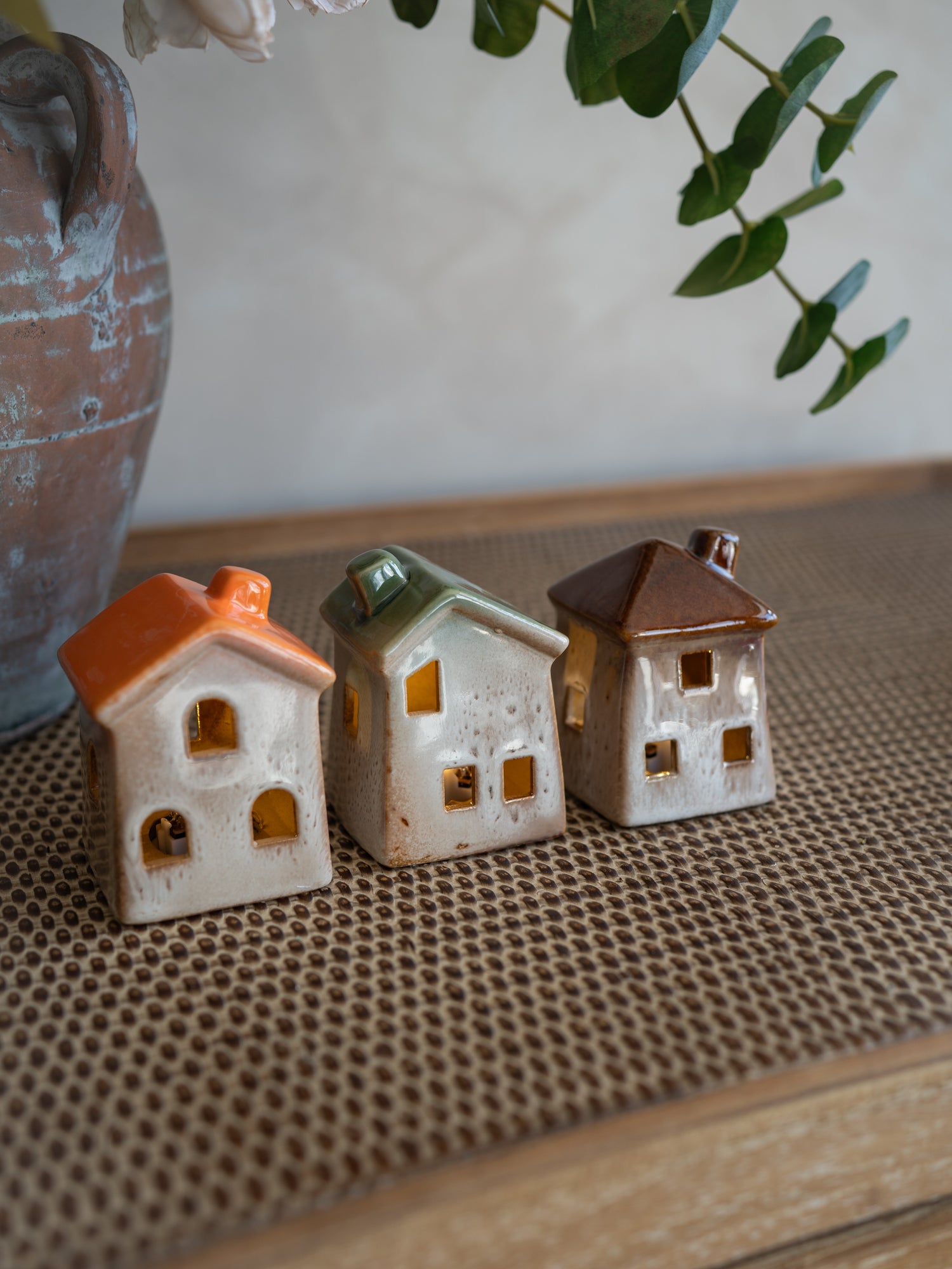 Three small ceramic house-shaped ornaments with orange, green, and brown roofs, displayed on a woven brown mat with a rustic vase and greenery in the background.