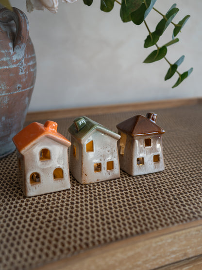 Three small ceramic house-shaped ornaments with orange, green, and brown roofs, displayed on a woven brown mat with a rustic vase and greenery in the background.