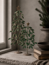 Two decorative metal Christmas tree ornaments with green leaves and gold star toppers, displayed on a stone windowsill next to a potted evergreen and stacked books.