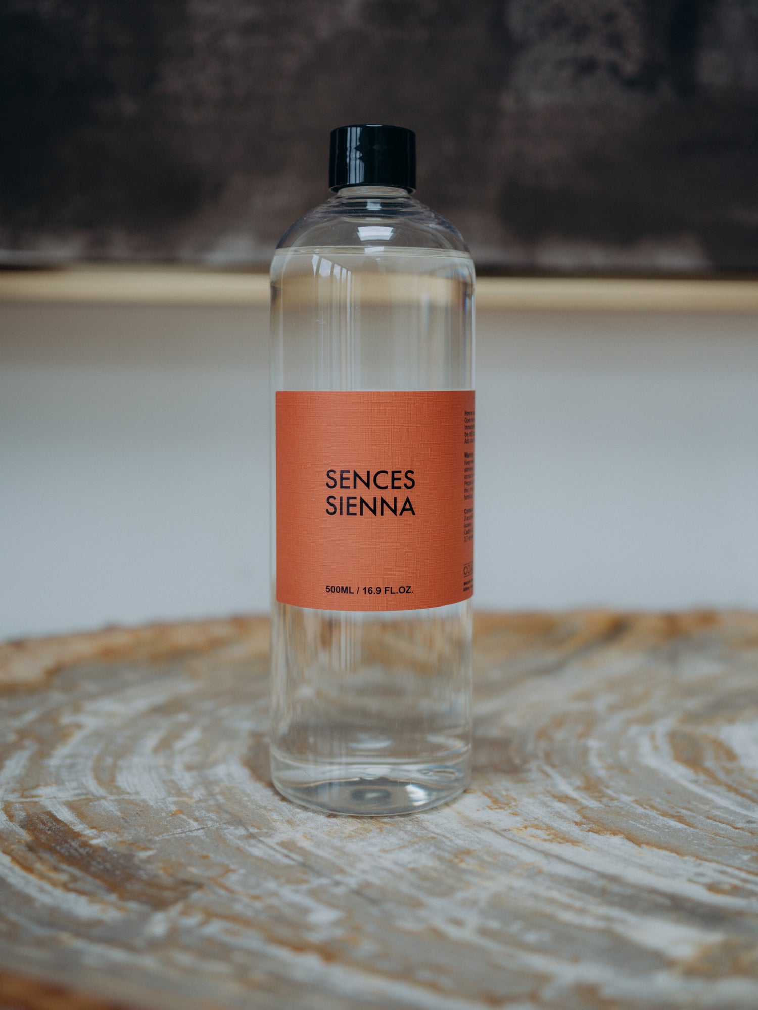 A clear plastic bottle of Sences Sienna diffuser refill oil with a black cap and a terracotta orange label, resting on a rustic wood slice coaster.