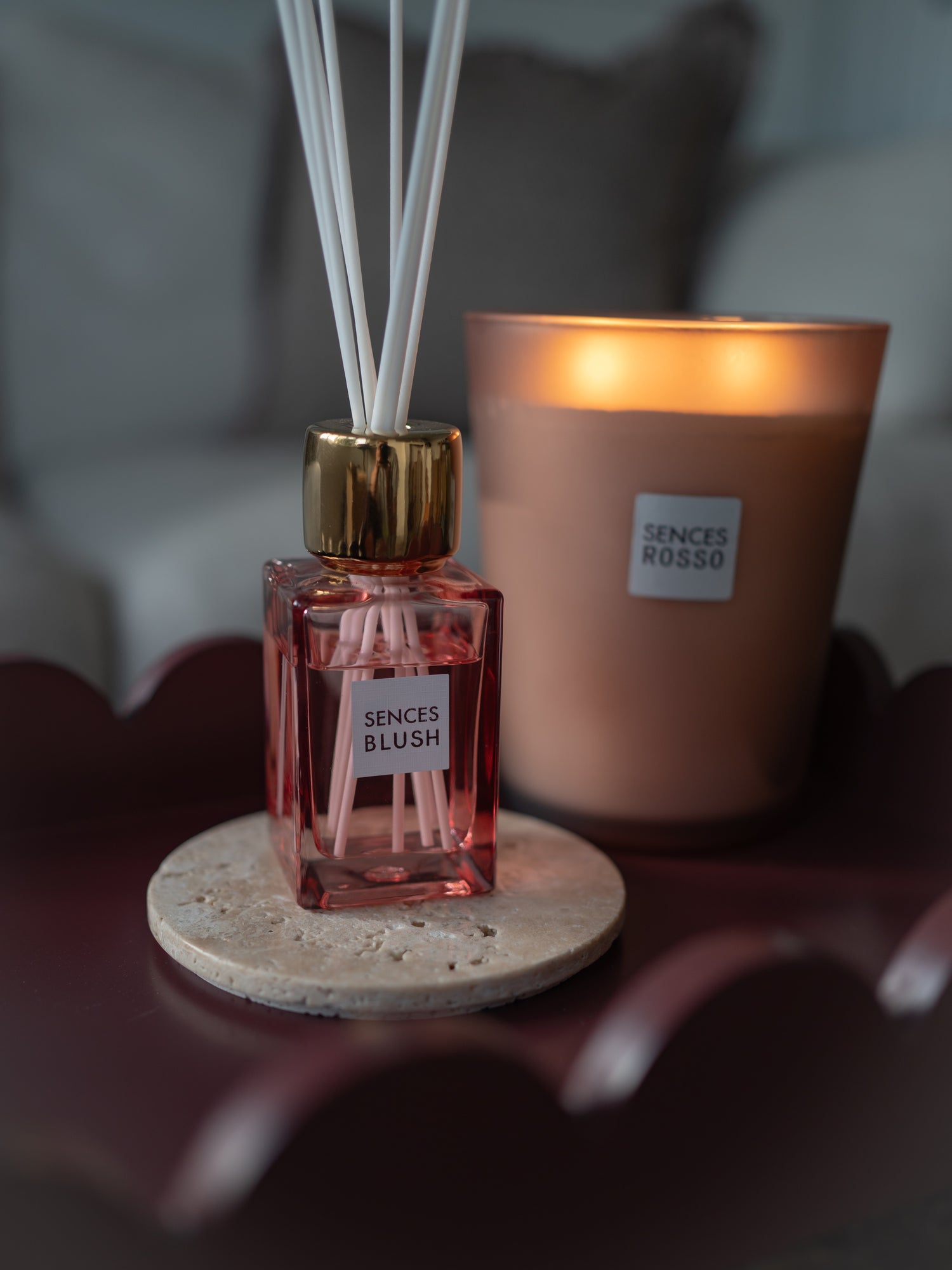 A blush sences reed diffuser bottle with pink liquid and white reeds sits on a stone coaster next to a lit candle labeled &
