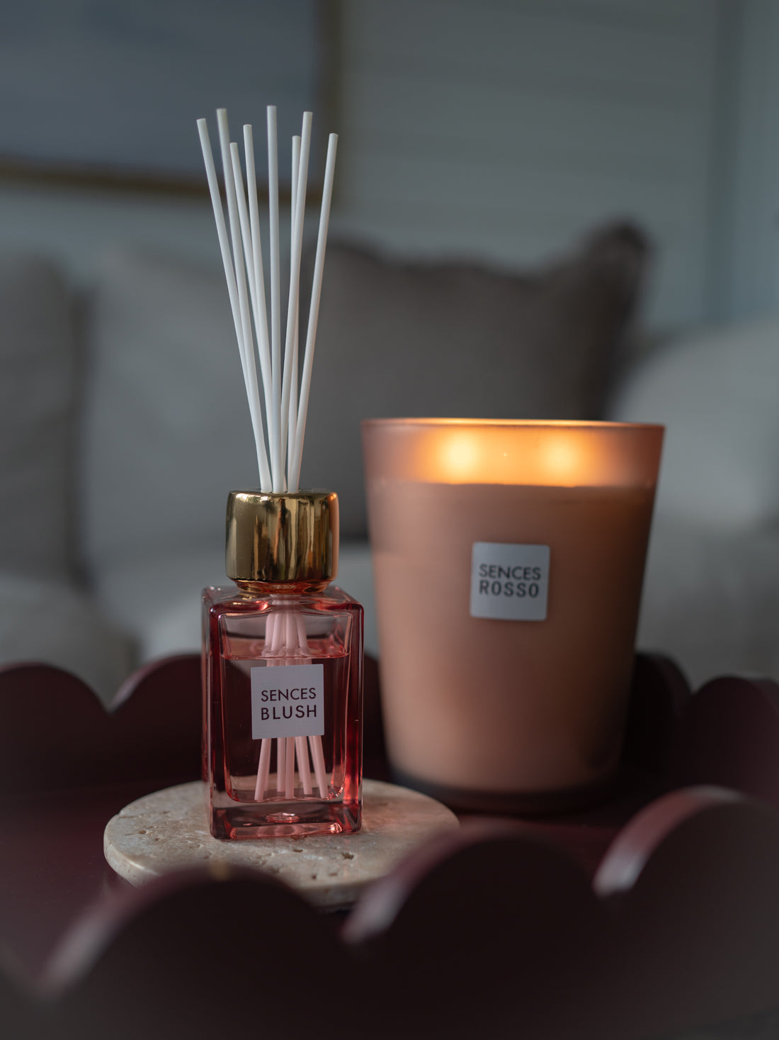 A reed diffuser bottle with white reeds and a gold cap stands beside a lit candle in a warm beige jar. Both containers have labels reading &