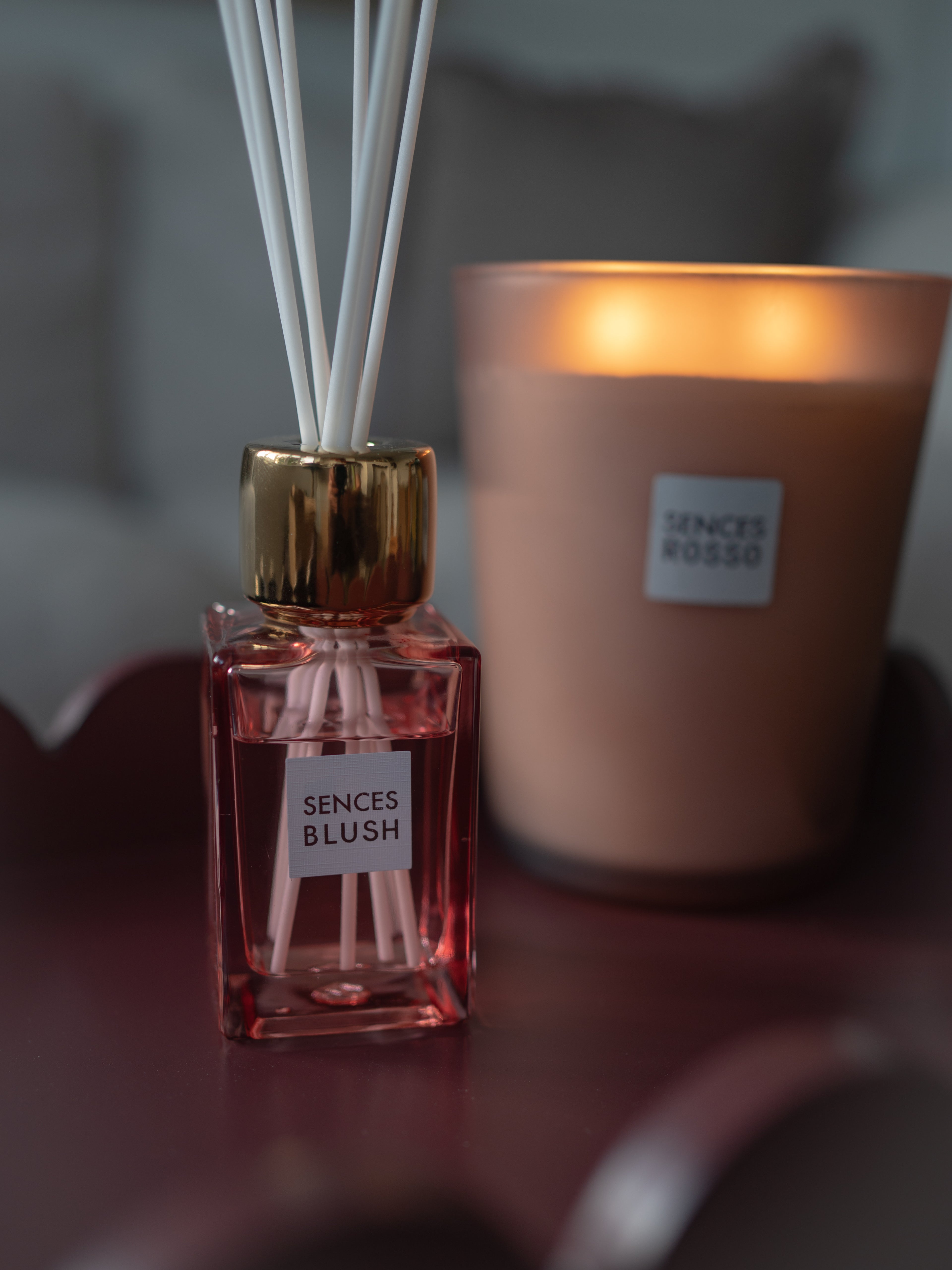 A Sences Blush reed diffuser bottle with pink liquid, a gold cap, and white reeds in the foreground, with a lit Sence&